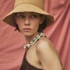 HAT ADIGE 1 Woman wearing the ADIGE handmade natural straw hat with a soft brim, portrait against a blush pink fabric backdrop.