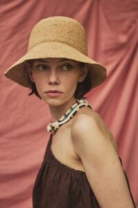 Woman wearing the ADIGE handmade natural straw hat with a soft brim, portrait against a blush pink fabric backdrop.