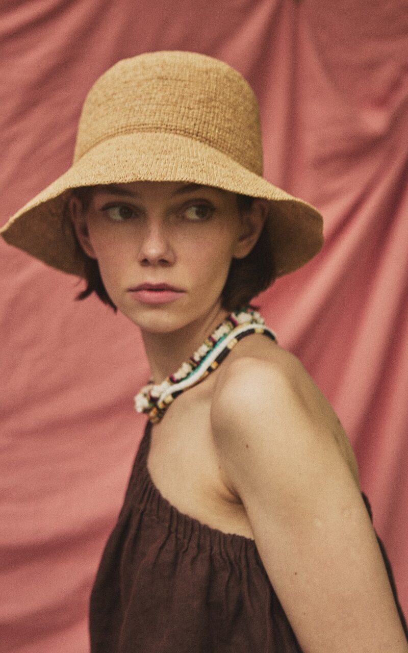Woman wearing the ADIGE handmade natural straw hat with a soft brim, portrait against a blush pink fabric backdrop.