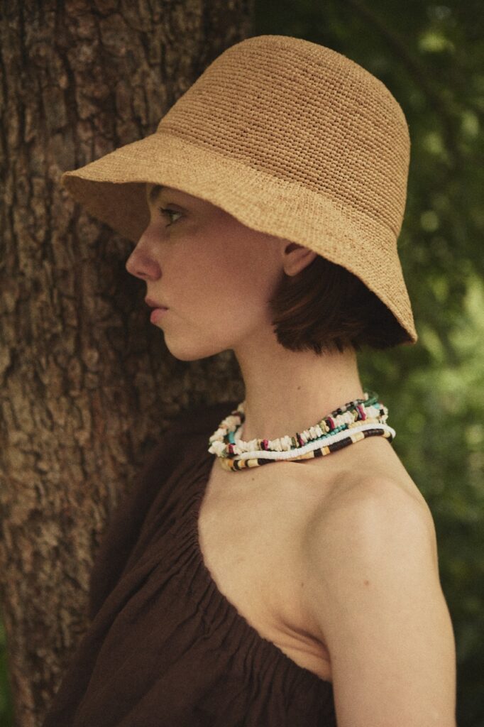 Side profile of a woman wearing the ADIGE handwoven natural straw hat outdoors beside a tree.