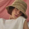 Artistic close-up of the TICINO handwoven sea grass hat creating patterned sunlight on a woman's face.
