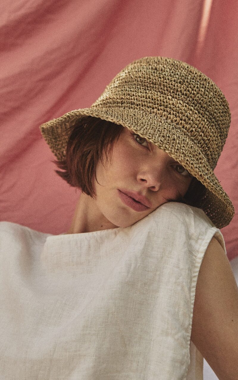 Artistic close-up of the TICINO handwoven sea grass hat creating patterned sunlight on a woman's face.
