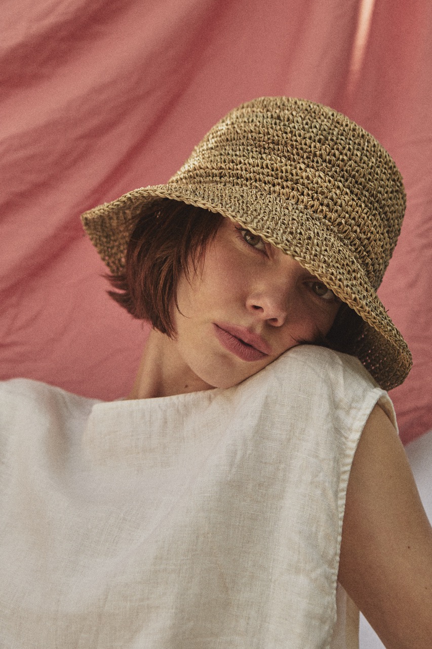 Artistic close-up of the TICINO handwoven sea grass hat creating patterned sunlight on a woman's face.